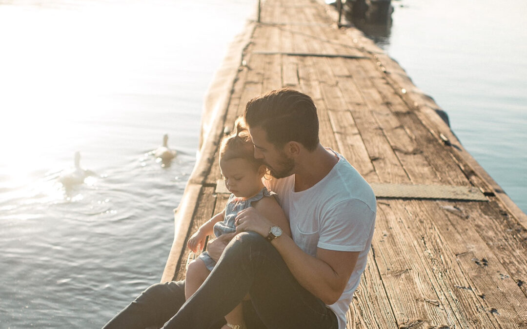 Father and daughter sitting on a dock next to water.